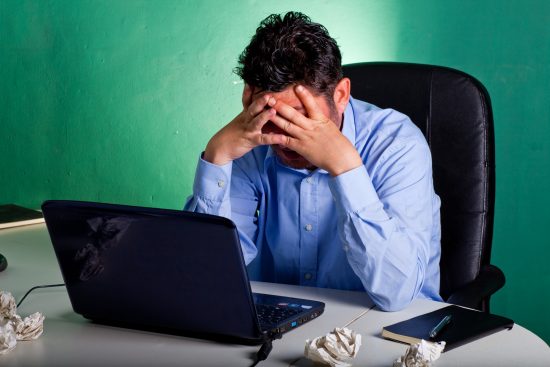 אדם עצוב, מחזיק ידיים על הפרצוף, מול מחשב נייד פתוח ודפים מקומטים